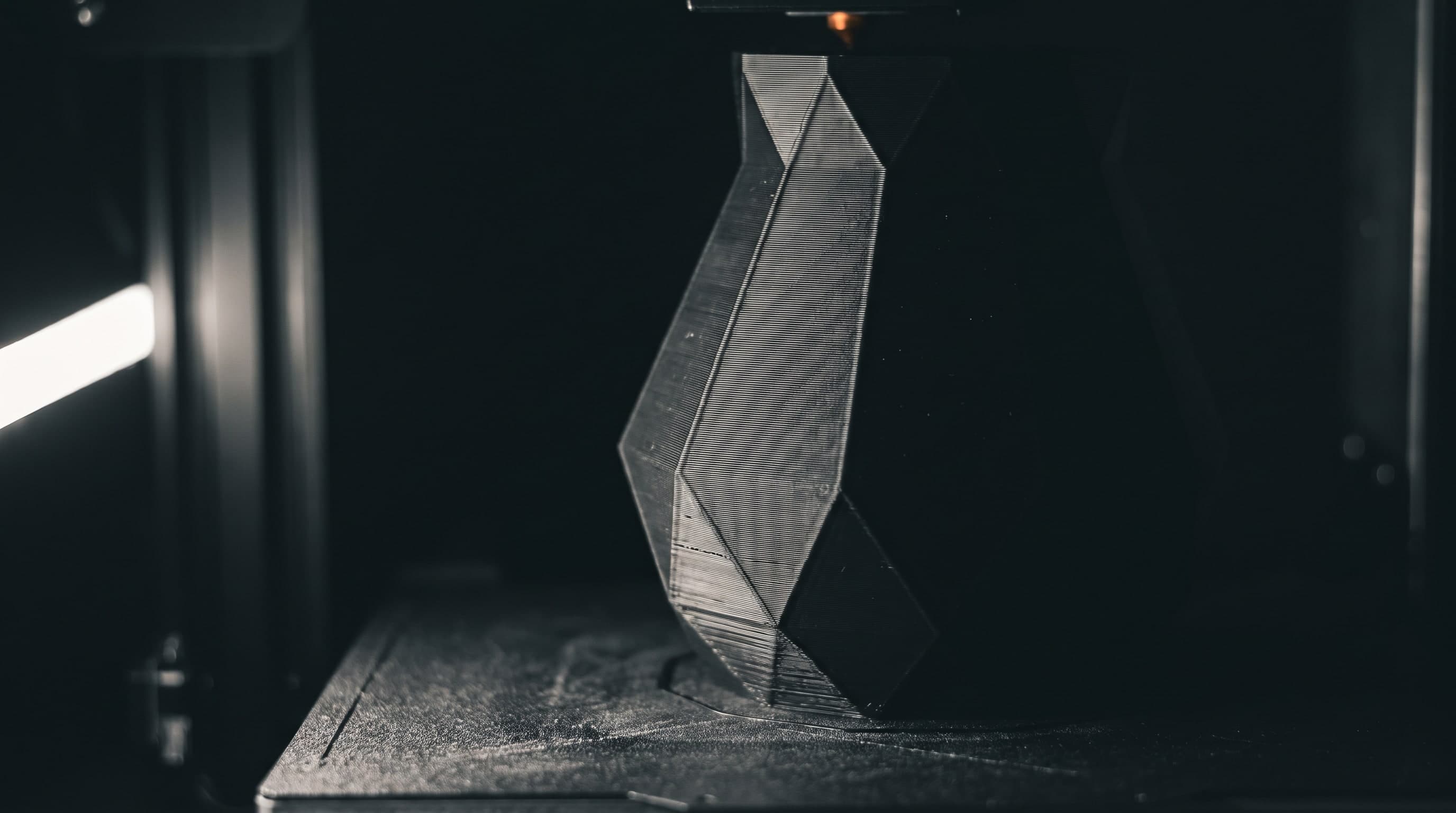 Kantiges, facettiertes graues Objekt auf dem Druckbett eines 3D-Druckers, oben glimmt die Düse